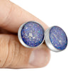 Close-up of a hand holding two sterling silver stud earrings featuring round blue and purple resin inlays with scattered silver flakes.