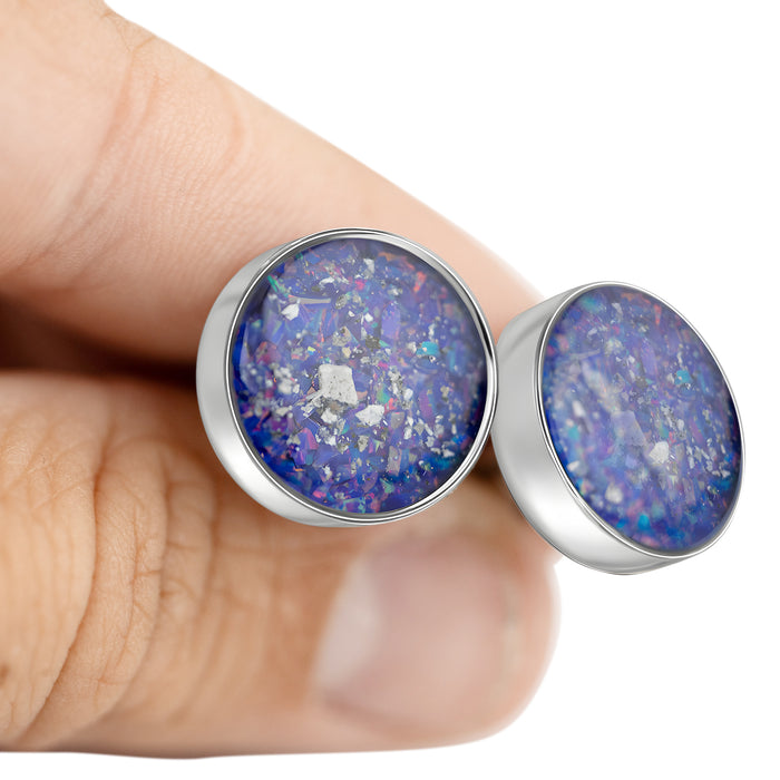 Close-up of a hand holding two sterling silver stud earrings featuring round blue and purple resin inlays with scattered silver flakes.