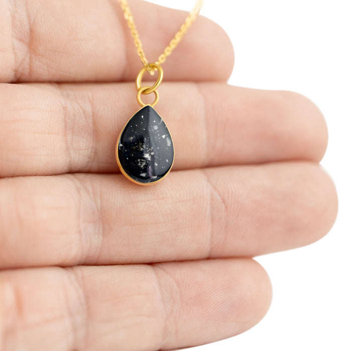 Close-up photo of a hand holding a gold-plated teardrop pendant with black resin and subtle silver flake accents.