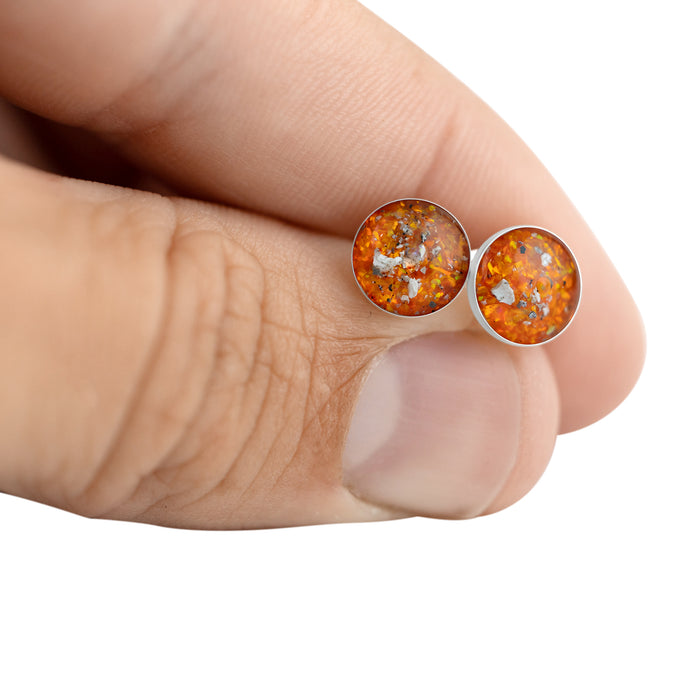 Close-up of a hand holding two sterling silver stud earrings featuring round vibrant orange and gold opal resin inlays.