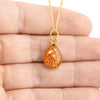 Close-up photo of a hand holding a gold-plated teardrop pendant with vibrant orange resin and internal gold and silver flakes.