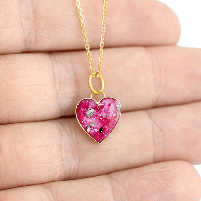 Close-up photo of a hand holding a gold-plated heart pendant with vibrant pink resin and shimmering silver flakes