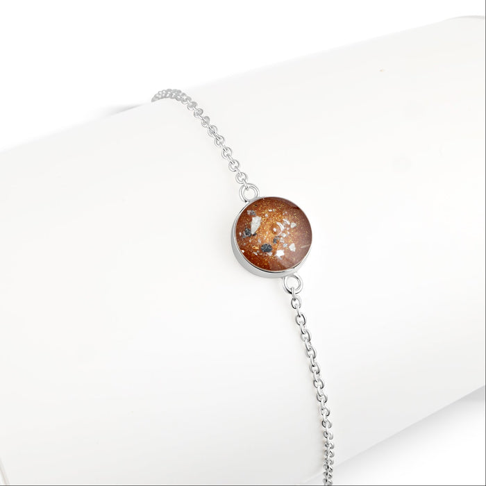 Thin sterling silver chain bracelet featuring a round charm with a brown resin inlay accented by gold and silver flakes.