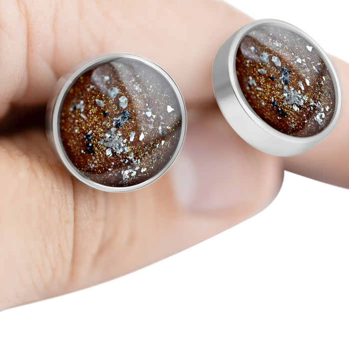 Close-up of a hand holding two sterling silver earrings featuring round bronze-brown resin inlays with gold and silver flakes.