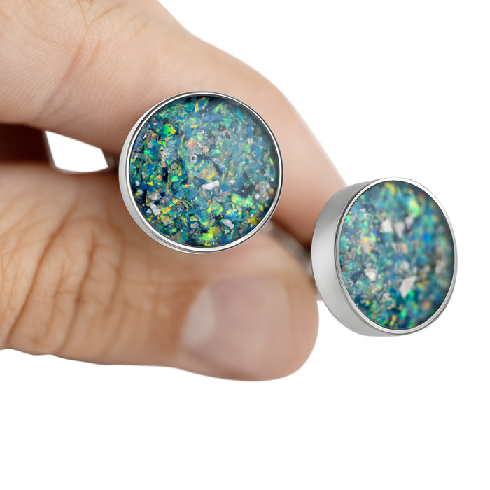 Close-up of a hand holding a pair of sterling silver stud cufflinks featuring round blue-green opal resin inlays.