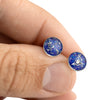 Close-up of a hand holding two sterling silver stud earrings featuring round blue and purple opal resin inlays.