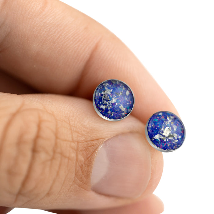 Close-up of a hand holding two sterling silver stud earrings featuring round blue and purple opal resin inlays.