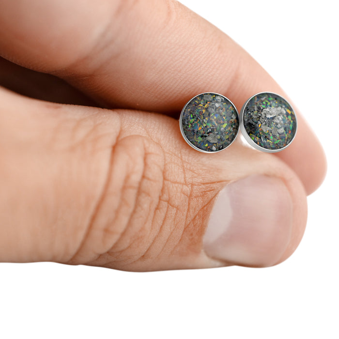 Close-up of a hand holding two sterling silver stud earrings featuring round dark grey and black opal resin inlays.