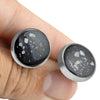 Close-up of a hand holding two sterling silver earrings featuring round black resin inlays with white and silver flakes.