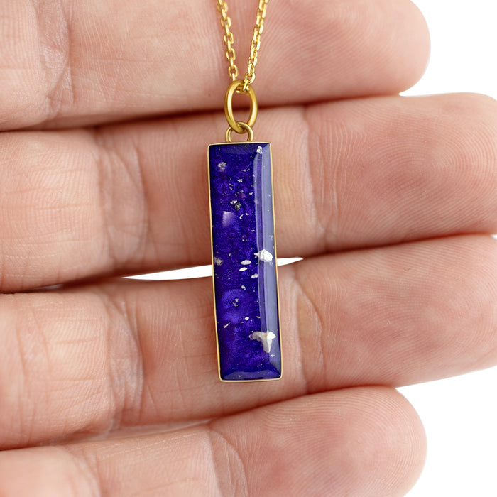 Hand holding a gold-plated bar pendant with vibrant blue resin and shimmering silver flakes.