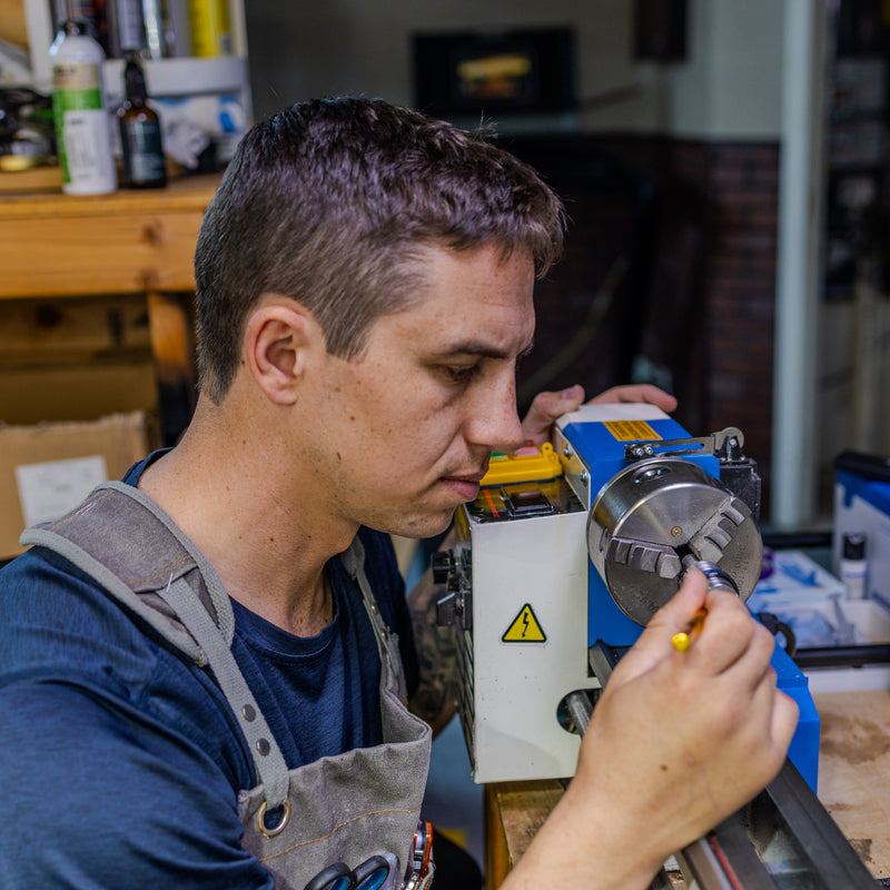 Close-up of a jeweler or artisan wearing an apron and working on a small lathe/precision machine in a workshop.
