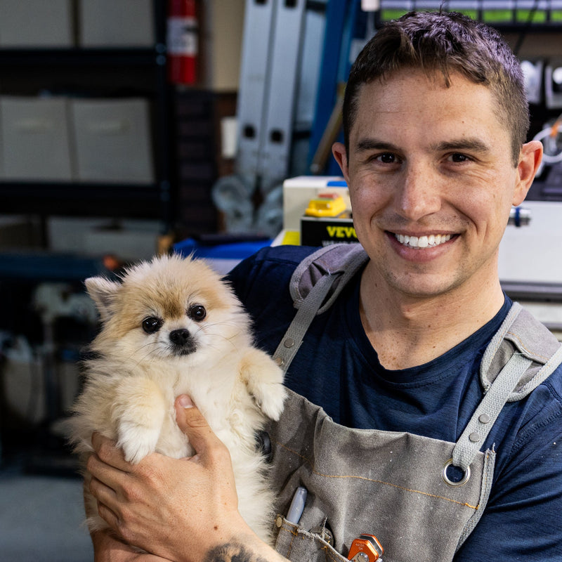 A smiling male artisan wearing a workshop apron, holding a fluffy light-colored Pomeranian dog.