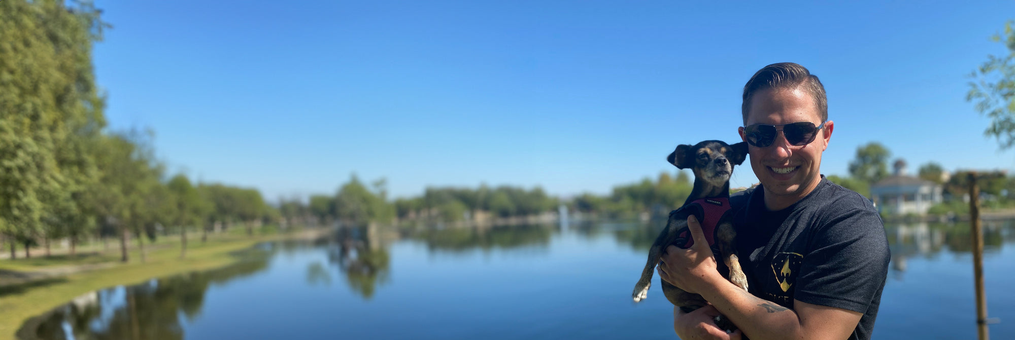 A smiling man holding a small dog in front of a blue lake on a sunny day.