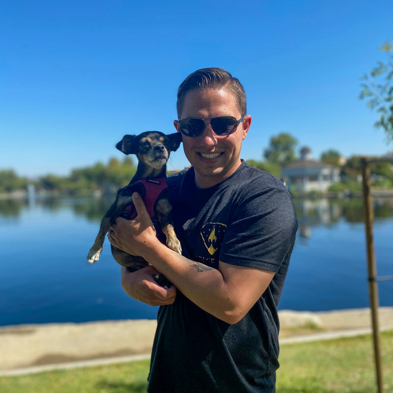 A smiling man wearing sunglasses and a black t-shirt, holding a small dog near a lake on a sunny day.